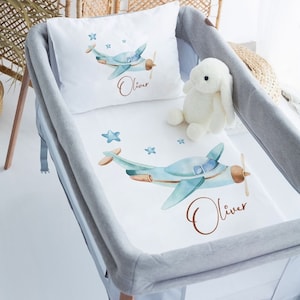 May include: A white crib bedding set with a blue airplane and stars design. The bedding set includes a fitted sheet, a pillowcase, and a plush bunny. The name "Oliver" is written on the bedding in brown cursive.