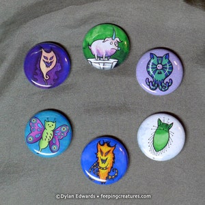 May include: Six colourful round buttons with different cartoon animal illustrations. The buttons feature a green pig with a horn, a green cat with a white background, a green butterfly with pink and blue wings, a purple cat with a white background, a blue cat with orange fur, and a purple cat with a white background.