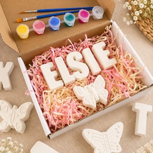 Könnte beinhalten: Ein Bastelset in einem Karton mit weißen Gipsbuchstaben, die ELSIE bilden, einem Schmetterling aus Gips, zwei Pinseln und einem Streifen mit sechs kleinen Farbtöpfen in Gelb, Pink, Blau, Grün, Magenta und Weiß, eingebettet in rosa Papier.