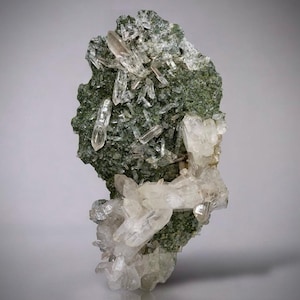 Puede incluir: Un espécimen mineral natural con una matriz verde oscuro cubierta de cristales de cuarzo transparente. Los cristales de cuarzo están agrupados y apuntan en varias direcciones, con algunas formaciones más grandes en la base. La apariencia general es áspera y texturizada.