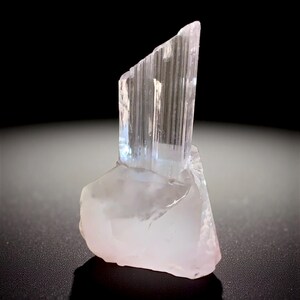 May include: A translucent, light pink crystal formation with a clear, vertical structure. The crystal has a rough, textured base and a smooth, angled top. The background is black, highlighting the crystal's natural beauty.