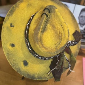 May include: A yellow felt cowboy hat with a black and white braided band and a brown scarf. The hat has a distressed look with black paint splatters and holes.