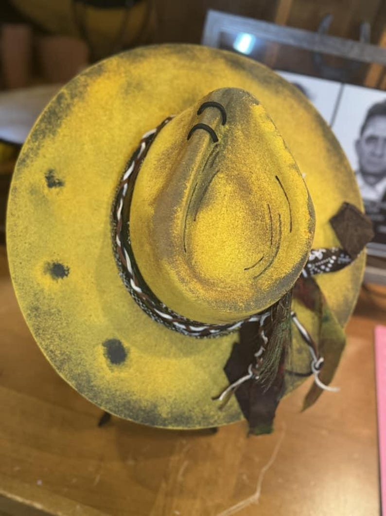 May include: A yellow felt cowboy hat with a black and white braided band and a brown scarf. The hat has a distressed look with black paint splatters and holes.
