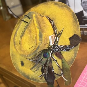 May include: A yellow felt cowboy hat with a brown bandana, a peacock feather, and a chain. The hat has a distressed look and is decorated with various embellishments.