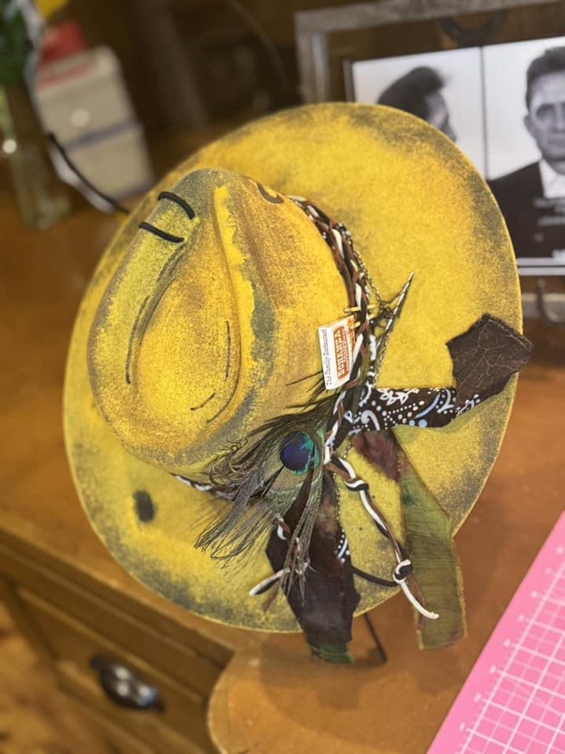May include: A yellow felt cowboy hat with a brown bandana, a peacock feather, and a chain. The hat has a distressed look and is decorated with various embellishments.