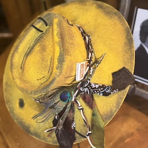 May include: A yellow cowboy hat with a brown and black bandana, a peacock feather, and a leather strap. The hat has a distressed look and is decorated with various embellishments.