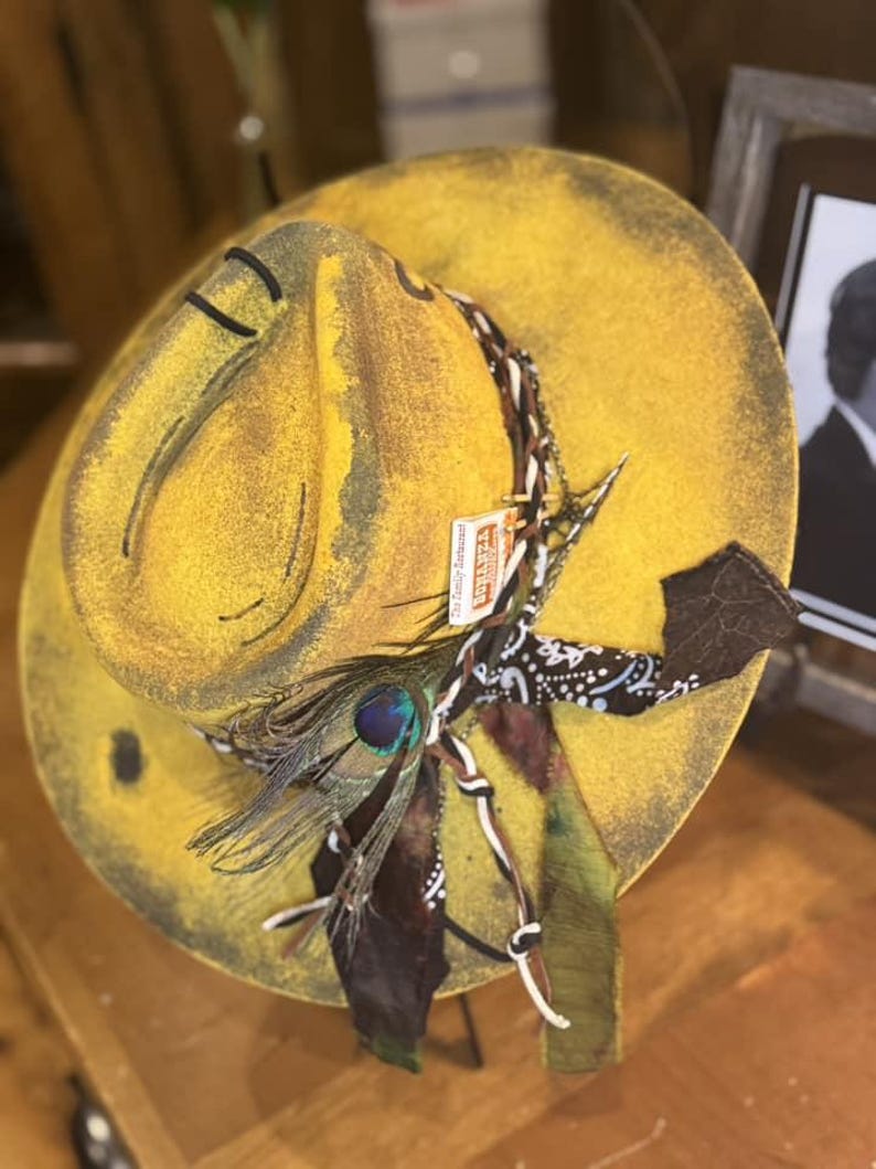 May include: A yellow cowboy hat with a brown and black bandana, a peacock feather, and a leather strap. The hat has a distressed look and is decorated with various embellishments.
