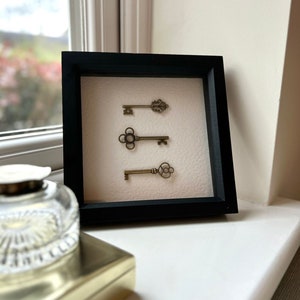 May include: A black framed picture with a white mat and three antique keys displayed on it. The keys are arranged vertically and are made of brass.