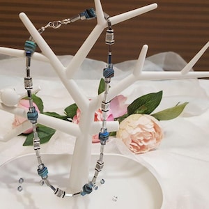 May include: A beaded necklace featuring a repeating pattern of metallic silver-toned cylindrical spacers and square-shaped beads in shades of blue, teal, and grey. The necklace is displayed on a white, tree-shaped jewellery stand with a circular base.