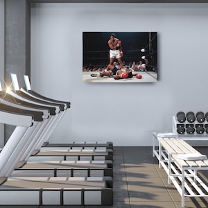 Peut inclure: Une photographie en noir et blanc d'un match de boxe. Le boxeur de gauche lance un coup de poing, tandis que le boxeur de droite est allong&eacute; sur le sol. La photographie est affich&eacute;e sur un mur dans une salle de sport avec des tapis roulants au premier plan.