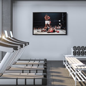Peut inclure: Une photo en noir et blanc d'un match de boxe. Le boxeur de gauche lance un coup de poing, tandis que le boxeur de droite est au sol. La photo est encadr&eacute;e et accroch&eacute;e au mur d'une salle de sport.