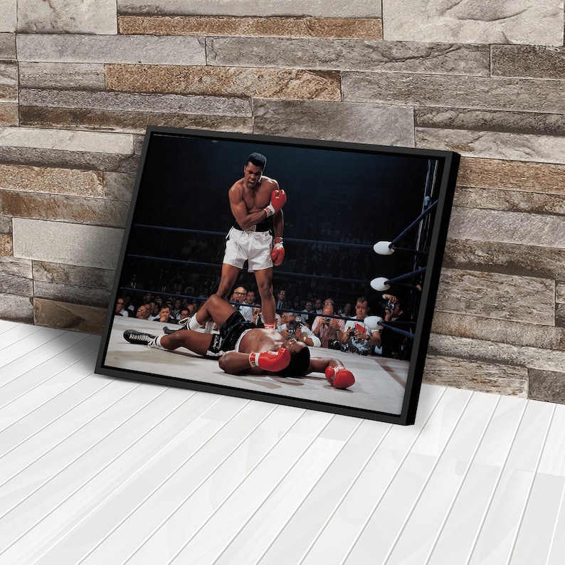 Peut inclure: Une photographie en noir et blanc d'un match de boxe. Un boxeur se tient au-dessus d'un autre boxeur qui est allong&eacute; au sol. Le boxeur debout porte un short blanc et des gants de boxe rouges. Le boxeur allong&eacute; au sol porte un short noir et des gants de boxe rouges. La photo est prise dans un ring de boxe avec une foule de personnes en arri&egrave;re-plan.