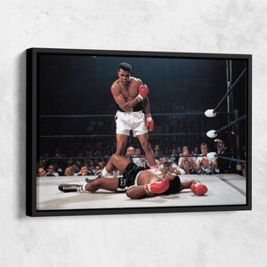 Peut inclure: Une photographie en noir et blanc d'un match de boxe. Le boxeur de gauche se tient au-dessus du boxeur de droite, qui est allong&eacute; au sol. Le boxeur de gauche porte un short blanc et des gants de boxe rouges. Le boxeur de droite porte un short noir et des gants de boxe rouges. La foule regarde le match en arri&egrave;re-plan.