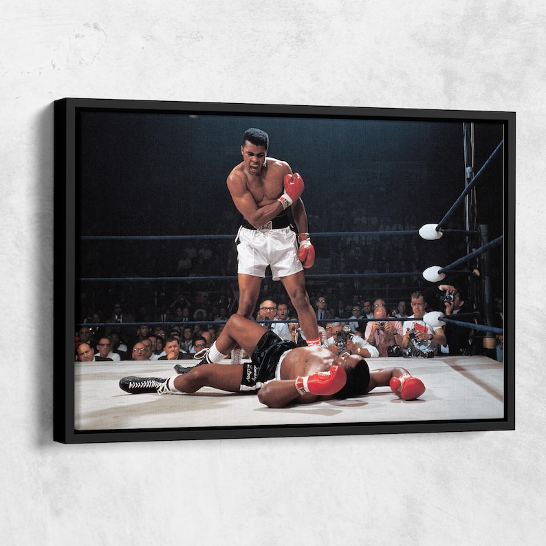 Peut inclure: Une photographie en noir et blanc d'un match de boxe. Le boxeur de gauche se tient au-dessus du boxeur de droite, qui est allong&eacute; au sol. Le boxeur de gauche porte un short blanc et des gants de boxe rouges. Le boxeur de droite porte un short noir et des gants de boxe rouges. La foule regarde le match en arri&egrave;re-plan.