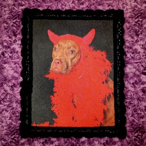 May include: A rectangular fabric patch featuring a brown dog wearing a red hood with pointed ears and a red feather boa. The image is set against a black background and is framed by a black crocheted border with a scalloped edge.