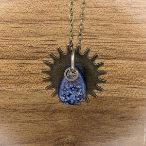 Puede incluir: Un collar con un colgante en forma de engranaje de color bronce y una piedra druzy azul en forma de lágrima en el centro. Una pequeña pieza de reloj metálica está unida a la parte superior de la piedra. El colgante cuelga de una cadena de metal.