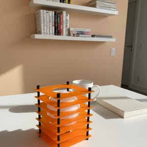 May include: An orange and black decorative lamp with a square design. The lamp has multiple orange square layers held together by black posts. A white mug and a book are on the white table next to the lamp. White shelves with books are in the background.