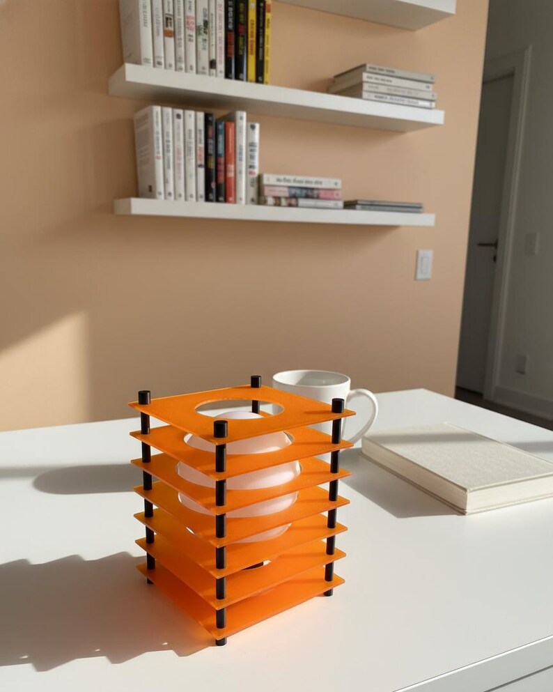May include: An orange and black decorative lamp with a square design. The lamp has multiple orange square layers held together by black posts. A white mug and a book are on the white table next to the lamp. White shelves with books are in the background.