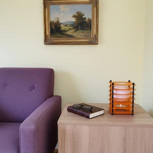 May include: A room interior featuring a purple armchair, a wooden side table, and a framed landscape painting. On the table are a closed book, eyeglasses, and an orange and black decorative lamp with a pink light.
