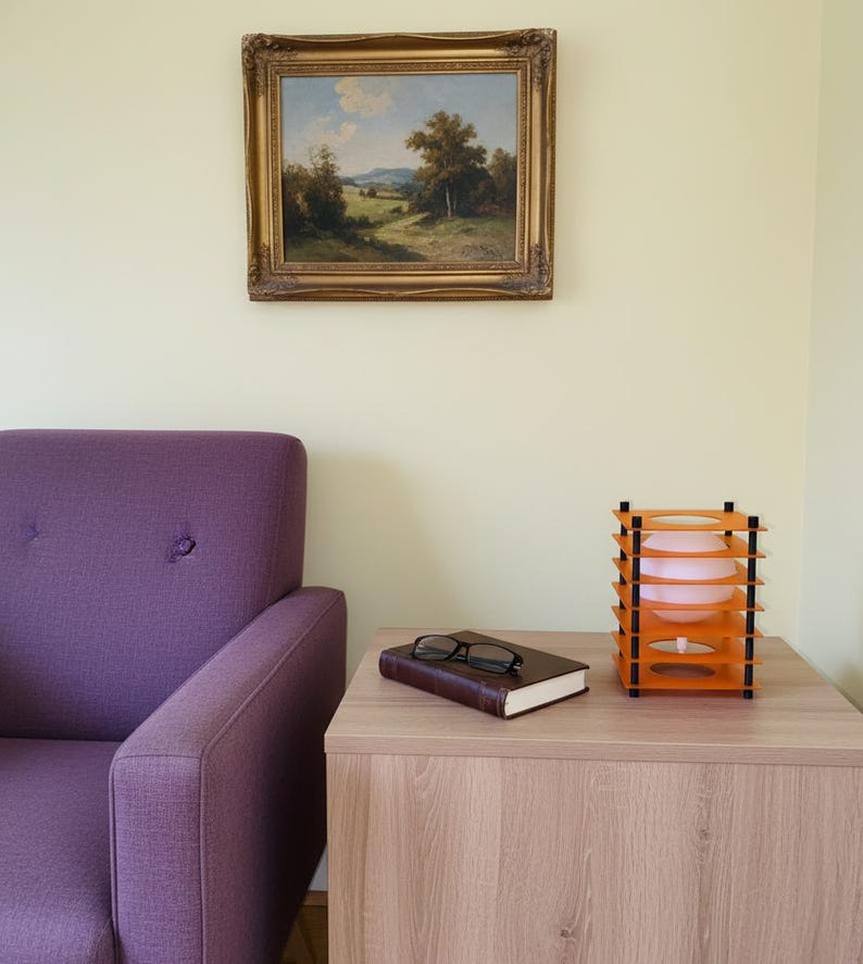 May include: A room interior featuring a purple armchair, a wooden side table, and a framed landscape painting. On the table are a closed book, eyeglasses, and an orange and black decorative lamp with a pink light.