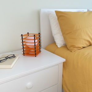 May include: An orange and black geometric table lamp with a pink bulb sits on a white nightstand. The lamp has a layered design. A book and eyeglasses are also on the nightstand. A bed with yellow bedding is in the background.