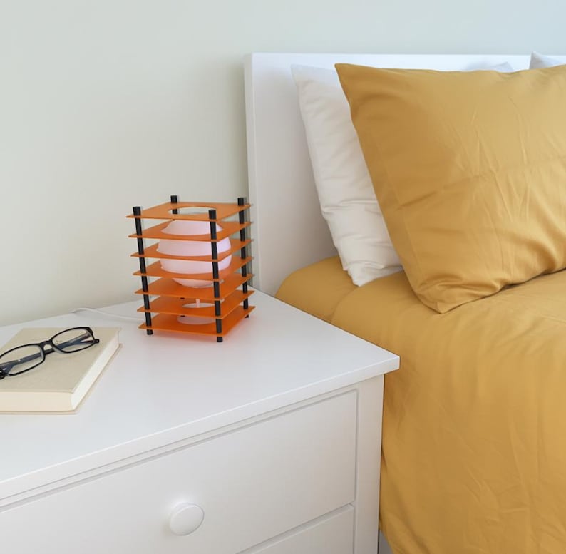 May include: An orange and black geometric table lamp with a pink bulb sits on a white nightstand. The lamp has a layered design. A book and eyeglasses are also on the nightstand. A bed with yellow bedding is in the background.