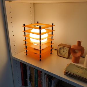 May include: An orange and black geometric table lamp with a spherical light source. The lamp is on a white shelf with a small brown clock, a decorative vase, and a stack of vintage postcards. Books line the shelf below.