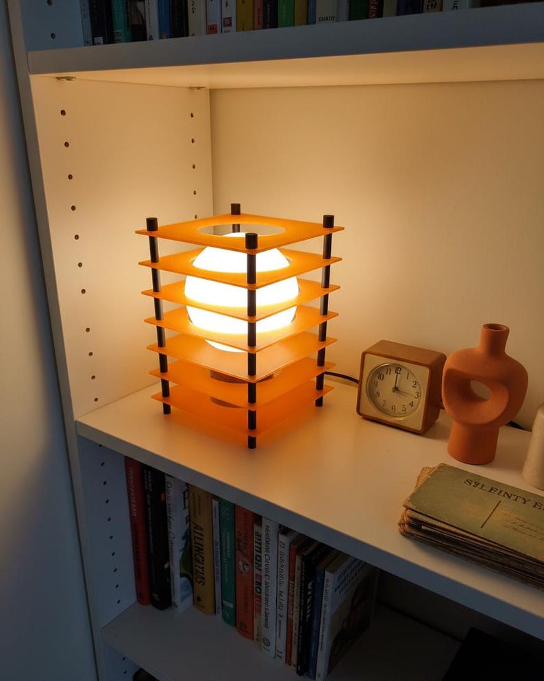 May include: An orange and black geometric table lamp with a spherical light source. The lamp is on a white shelf with a small brown clock, a decorative vase, and a stack of vintage postcards. Books line the shelf below.