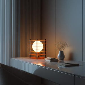 May include: A modern table lamp with a spherical light source encased in a wooden frame. The lamp sits on a white desk next to a small vase with dried flowers and a stack of books. The lamp's warm glow illuminates the room.