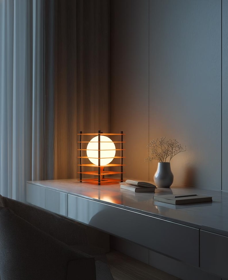 May include: A modern table lamp with a spherical light source encased in a wooden frame. The lamp sits on a white desk next to a small vase with dried flowers and a stack of books. The lamp's warm glow illuminates the room.