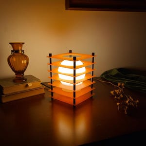 May include: A modern orange and black cube lamp with a white globe light inside. The lamp is on a dark wooden surface, next to a brown vase and a stack of books. The lamp's design features layered square panels.
