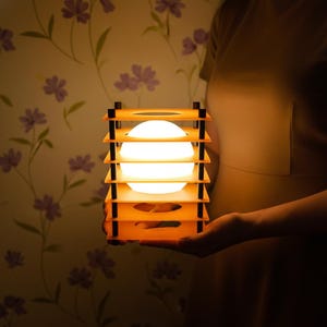May include: A glowing, spherical light fixture encased in a layered, orange wooden frame with black accents. The lamp is held in front of a floral wallpaper background, creating a warm, inviting ambiance. The light source is a bright, white orb.