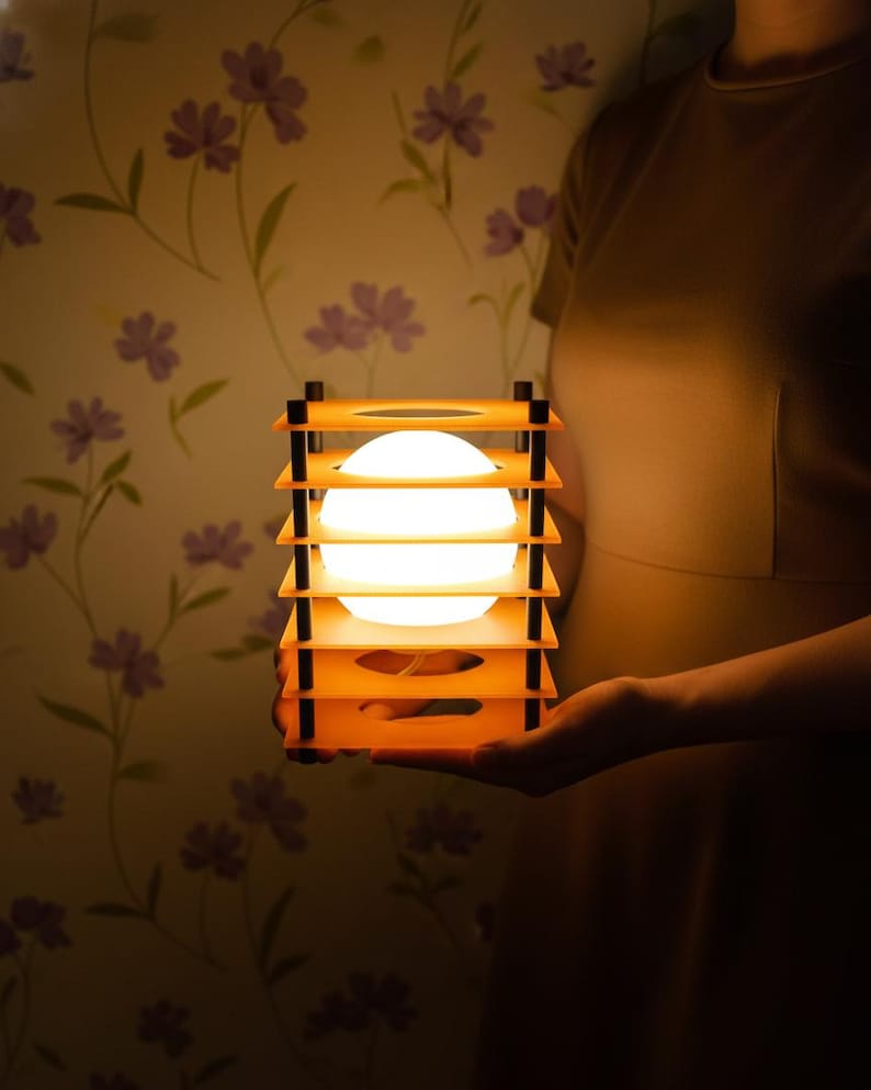 May include: A glowing, spherical light fixture encased in a layered, orange wooden frame with black accents. The lamp is held in front of a floral wallpaper background, creating a warm, inviting ambiance. The light source is a bright, white orb.