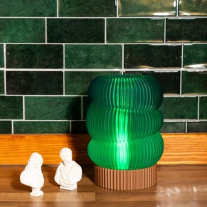 May include: A green glass table lamp with a wooden base. The lamp has a unique, layered design with a ribbed texture. The lamp is sitting on a wooden surface.