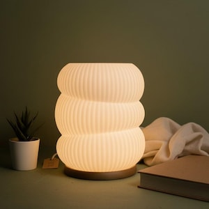 Può includere: Una lampada da tavolo color crema con un design impilato e arrotondato. La lampada presenta dettagli a coste verticali e poggia su una base in legno. Una piccola pianta in vaso e un libro chiuso sono accanto alla lampada.
