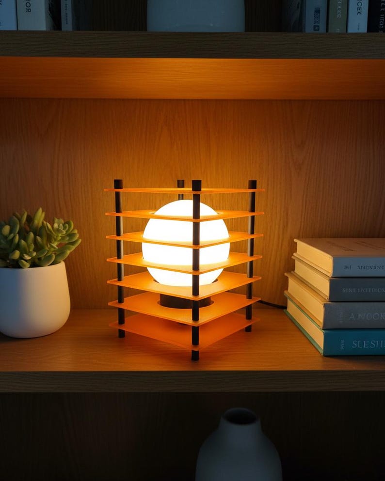 May include: A modern orange and black table lamp with a spherical light source. The lamp has a unique geometric design with multiple square shelves. The lamp is on a wooden shelf with books and a potted plant.