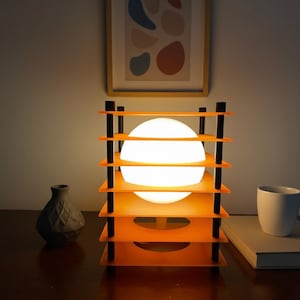 May include: A modern orange and black table lamp with a spherical white light source. The lamp has a tiered design with horizontal orange shelves and vertical black supports. A framed abstract art print hangs in the background.