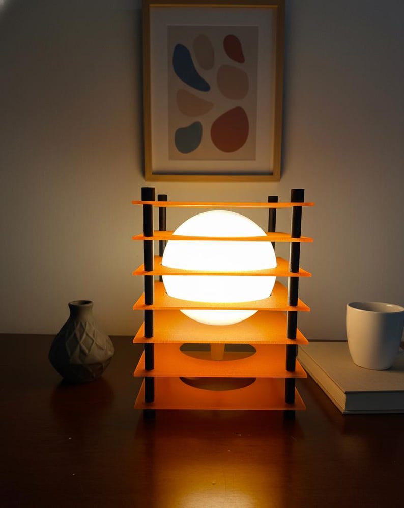 May include: A modern orange and black table lamp with a spherical white light source. The lamp has a tiered design with horizontal orange shelves and vertical black supports. A framed abstract art print hangs in the background.