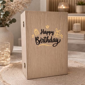 Puede incluir: Una caja de madera marrón claro con las palabras "Happy Birthday" en letras negras y doradas. La caja tiene una superficie texturizada y está decorada con ilustraciones de cajas de regalo. La caja está sobre un salvamanteles beige tejido.