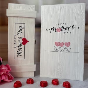 May include: A white wooden gift box and card set for Mother's Day. The box has the text "Happy Mother's Day" with a heart. The card features the same text and three pink heart-shaped flowers. Red heart-shaped candies are in front.