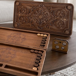 Op de afbeelding: Een houten backgammonspel met een open bord, lichte en donkere ronde schijven, twee blauwe dobbelstenen en een bijpassend rechtopstaand deksel met gedetailleerd bloemmotief. Een kleine houten houder voor de dobbelstenen staat ernaast.