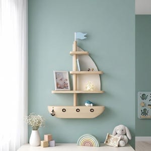 Puede incluir: Una estantería de pared de madera con forma de velero, que cuenta con tres niveles horizontales de almacenamiento, un mástil vertical, una bandera azul claro en la parte superior y una base de casco curvada con tres ojos de buey y un ancla.