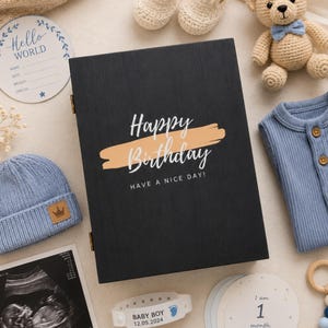 Pode incluir: Uma caixa de madeira preta com o texto Happy Birthday e Have a Nice Day impresso na frente. Os itens ao redor incluem um urso de peluche de crochê, sapatinhos, um gorro azul, um body azul, uma ecografia e cartões de marcos do bebê.