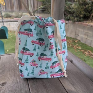 May include: A light blue drawstring bag featuring a pattern of pink trucks with Christmas trees and green fir trees. It has a cream drawstring closure. The bag is on a wooden surface.