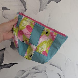 May include: A handmade, zippered pouch with a tropical flamingo pattern. The pouch features a pink zipper, teal blue panels, and a patchwork design with pink flamingos and green palm leaves on a black background.