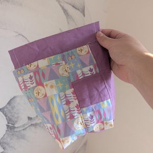 May include: A purple fabric bag with a patterned front pocket. The pocket features a colorful design with skeleton figures, triangles, and floral elements. The bag is held by a hand, showcasing the contrast between the solid purple and the patterned fabric.