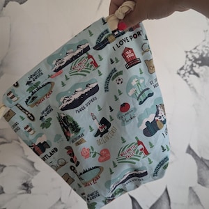 Peut inclure: Un sac bleu clair avec cordon de serrage, orné d'un motif de carte avec des illustrations de monuments de l'Oregon, dont Crater Lake et la Willamette Valley, et le texte "I Love Portland". Le sac a un cordon de serrage crème.