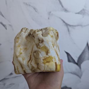 May include: A small, square, drawstring pouch with a marbled pattern in shades of cream, brown, and yellow. The fabric is gathered at the top, creating a textured, organic appearance. The pouch is held in a hand.