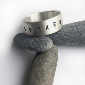 May include: A silver ring with the word "SPOKEN" stamped on the outside. The ring is resting on top of several gray stones. The ring has a wide band and a brushed metal finish.
