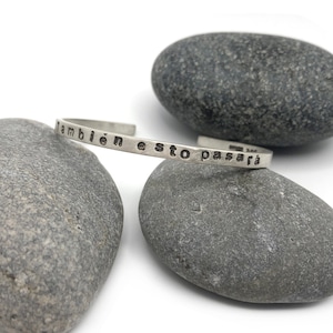 May include: A silver cuff bracelet with the stamped phrase "también esto pasará" is displayed. The bracelet rests on two gray stones against a white background. The bracelet is a simple, elegant design.